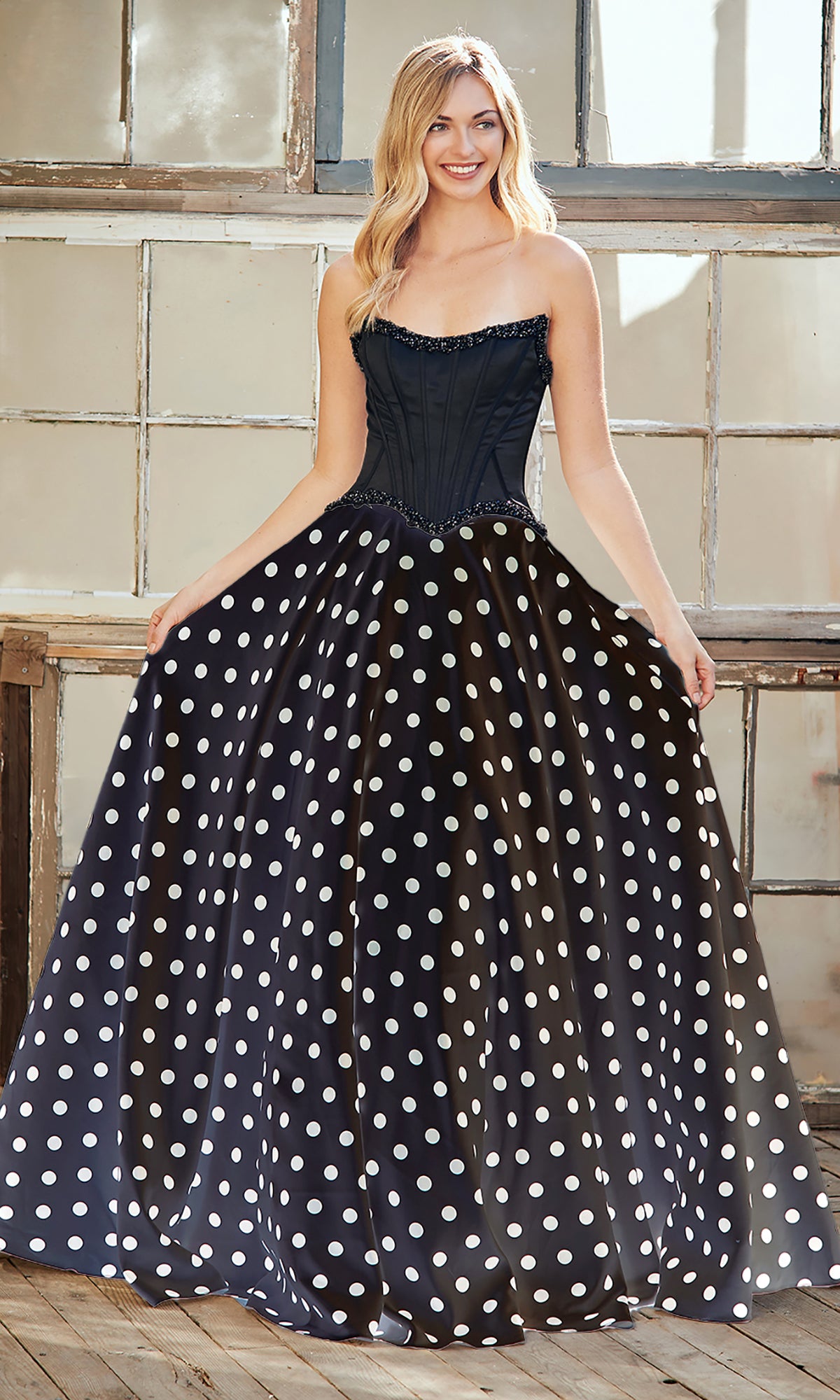 Indoors, a woman wears the Amelia Couture 8071 two-piece strapless prom gown featuring a fitted bodice and a full skirt with white polka dots.